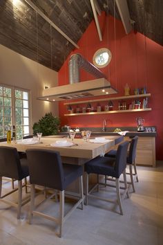 a dining room table surrounded by chairs in front of an open kitchen area with red walls