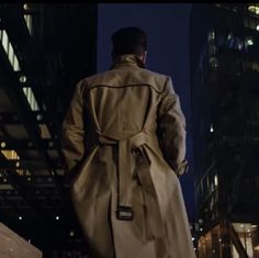 a man in a trench coat is walking down the street at night with his back to the camera