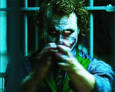 the joker is holding his finger up in front of jail cell with bars behind him