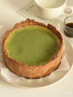 a green cake on a white plate next to a cup of coffee and saucer