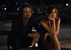 a man sitting next to a woman on top of a sidewalk at night with headphones in her ears