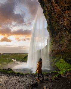 a person standing in front of a waterfall with water coming out of the side and green grass on the other side