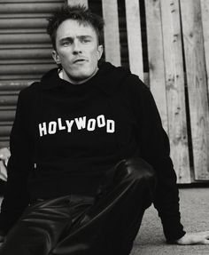 a man sitting on the ground in front of a wooden fence wearing a hollywood hoodie