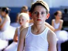 a young boy in a white tank top is surrounded by ballet dancers