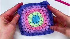 two hands are holding a crocheted square in front of a pink and blue object