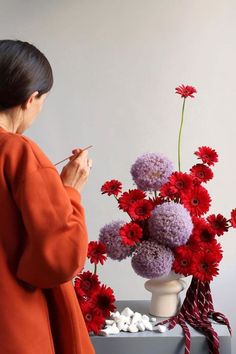 a woman in an orange coat painting flowers on a vase with white rocks and red carnations