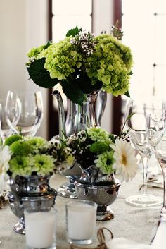 a table topped with silver vases filled with green and white flowers next to wine glasses