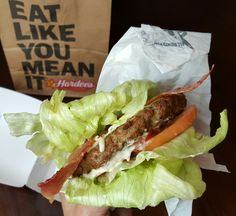 a hand holding a sandwich with lettuce, tomato and meat on it in front of a paper bag