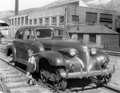 a black and white photo of a boy standing next to an old car on train tracks