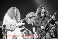 two men playing guitars on stage at a music festival, one with long hair and the other with an electric guitar