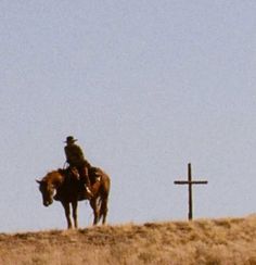 a man riding on the back of a brown horse next to a cross in a field