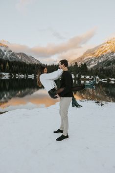 a man and woman standing in the snow by a lake with mountains in the background