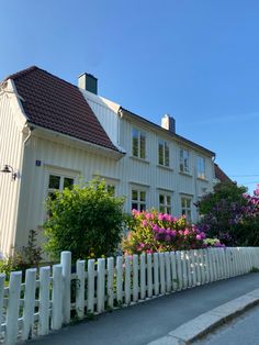 a white picket fence is in front of a house with pink and purple flowers on it