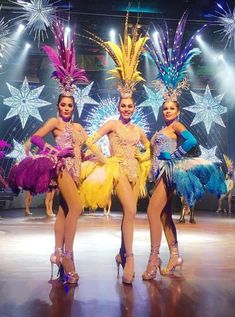 three women in brightly colored costumes standing on stage with fireworks and stars behind them, all wearing high heels