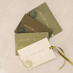 three different colored envelopes with ribbons tied around them on a white tablecloth, one is green and the other has gold foil