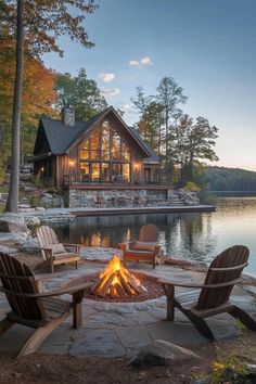 a fire pit in front of a lake with chairs around it and a log cabin on the other side