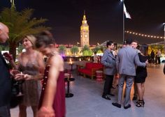 people are standing around talking and drinking at an outdoor event in front of a clock tower