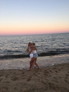 two women hug each other on the beach as the sun sets in the distance behind them