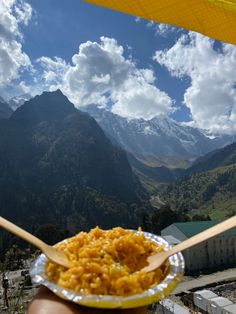 a person holding a spoon with food in front of some mountains and clouds on a sunny day