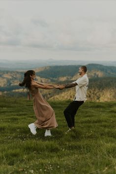 a man and woman holding hands while standing on top of a lush green field with mountains in the background