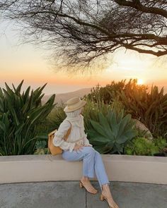 a woman sitting on top of a cement bench next to a lush green forest at sunset