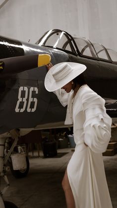 a woman in a white dress and hat standing next to an airplane