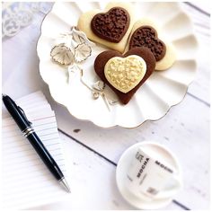 three heart shaped cookies on a plate next to a cup of coffee and a pen