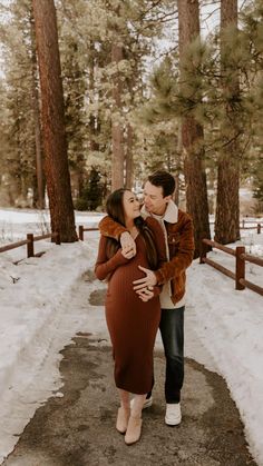 a man and woman standing in the snow next to trees with their arms around each other