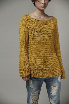 a woman with short hair wearing a yellow sweater and ripped jeans, standing in front of a gray background