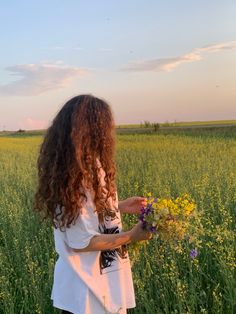 a woman standing in a field holding flowers and looking at the sun setting behind her