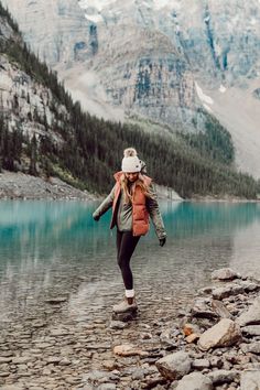 a woman walking across a rocky beach next to a lake with mountains in the background