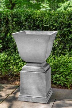 a cement urn sitting on top of a sidewalk next to some bushes and trees