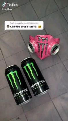 two monster energy drink cans sitting on the ground next to a pink and black can