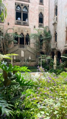 an outdoor garden with lots of plants and flowers in front of a large pink building