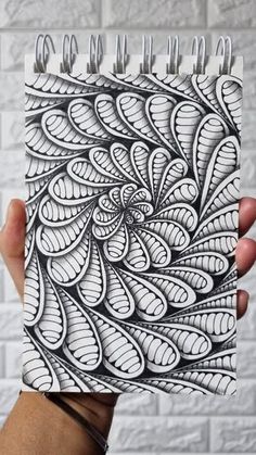 a hand holding up a spiral notebook with black and white designs on the cover, in front of a brick wall