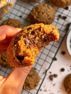 a person holding up a cookie in front of some chocolate chip cookies on a cooling rack