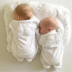 two babies laying next to each other on top of a bed with white sheets and pillows