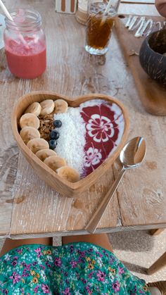 a wooden table topped with a heart shaped bowl filled with fruit and cereal next to a woman's legs