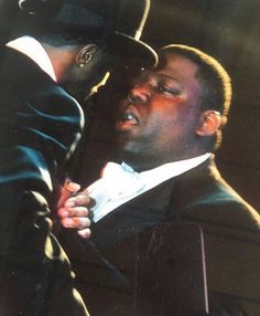 a man in a suit and top hat is hugging another man's neck with his eyes closed