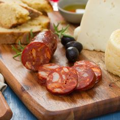 a wooden cutting board topped with sliced up sausages next to bread and olives