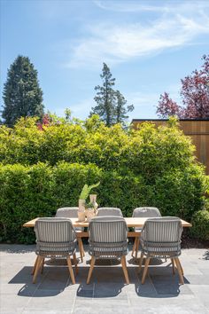 an outdoor dining table with four chairs in front of some bushes and trees on a sunny day