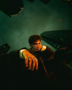 a man holding his hand up to his face while sitting in front of some tall buildings