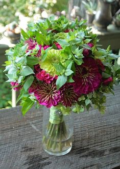 a vase filled with purple and green flowers on top of a wooden table next to trees