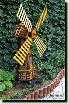 a toy windmill made out of wood sitting in front of some plants and bushes on the ground