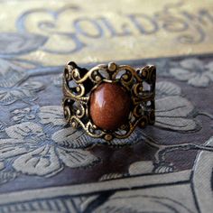 10x8mm brown goldstone cabochon on an antiqued brass adjustable filigree ring. Easy to adjust to any size. Shade of brownish orange may vary slightly if at all. About the stone: This is a man made gemstone. Thousands of tiny copper particles glitter against a brown background. The story has it that a monk accidentally dropped copper shavings into a vat of molten glass to create this material. Antique Adjustable Bronze Rings, Adjustable Bronze Rings With Antique Finish, Adjustable Oval Brown Rings, Adjustable Oval Rings In Brown, Vintage Adjustable Filigree Ring Gift, Vintage Adjustable Amber Ring, Adjustable Antique Brown Jewelry, Antique Adjustable Brown Jewelry, Stone Wedding Rings