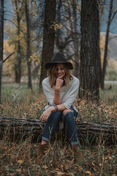 a woman wearing a hat sitting on a log in the woods with her hand under her chin