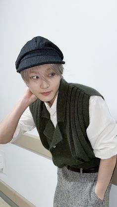 a young man wearing a green vest and hat with his hand on his head while leaning against a wall