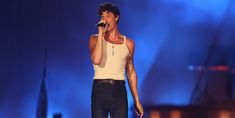 a male in a white tank top and black jeans singing into a microphone on stage