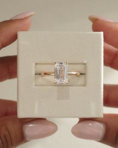 two hands holding an engagement ring with a diamond in the middle and a white box behind it