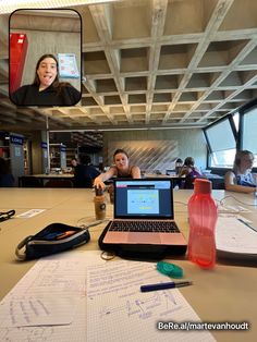 two women sitting at a table with laptops and notebooks on it, talking to each other